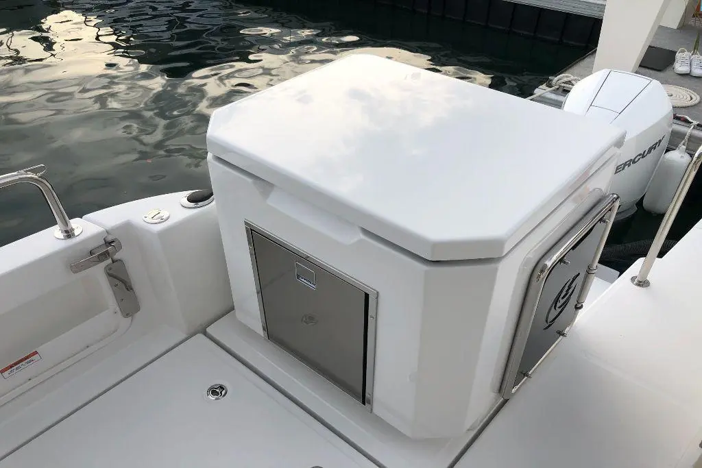 A white boat cooler with a metal door sits on the deck, against a backdrop of water and a motorboat.